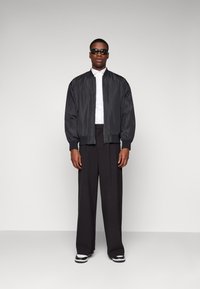 Giacca bomber nera con polsini a costine, indossata sopra una camicia bianca a bottoni, abbinata a pantaloni larghi neri e sneakers bianco e nere.