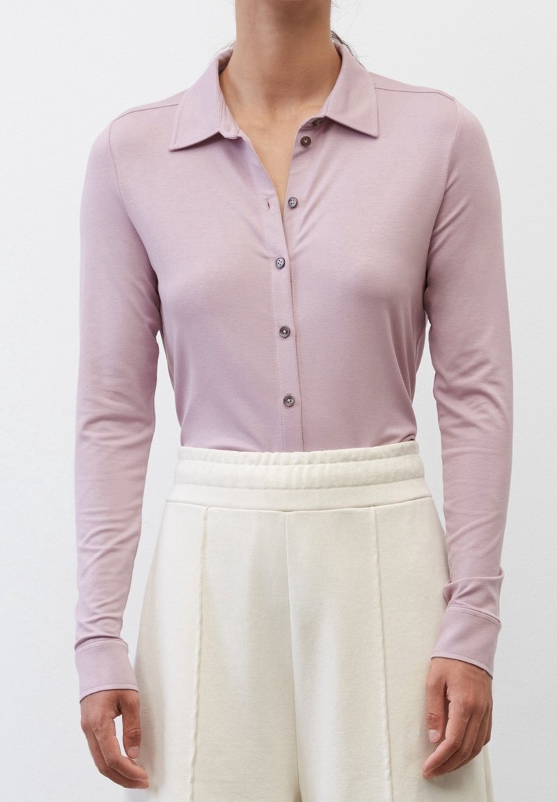 Femme portant une chemise à manches longues violet clair boutonnée, rentrée dans un pantalon taille haute plissé de couleur blanc cassé.