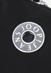 Črn najlonski material z okroglim srebrnim logotipom, okoli katerega je besedilo "JOOP! JEANS" in osrednjim izrezom. Besedilo je poudarjeno.