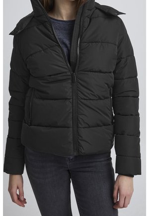 Femme portant une veste matelassée noire zippée par-dessus une chemise bleu foncé et un jean sombre sur un fond neutre.
