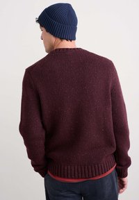 Burgundijas trikotāžas džemperis ar strukturētu apdari un ribotu apkakli un piedurknēm, apvienots ar tumši zilu parūku un sarkanu kreklu zem tā.