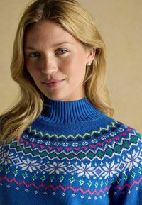 Zils adīts džemperis ar teksturētu augstu apkakli. Satur krāsainus ģeometriskus rakstus un sniega pārslu motīvus rozā, zaļā un baltā krāsā.