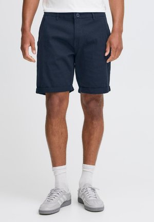 Shorts din bumbac bleumarin cu închidere cu nasture, manșete răsucite și buzunare laterale, asortați cu adidași gri și șosete albe până la glezne.