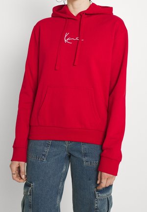 Person wearing a red hooded sweatshirt with front pocket and drawstrings, paired with blue denim cargo jeans.