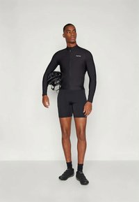 Μαύρη ποδηλατική στολή με μακριά μανίκια και φερμουάρ μπροστά, σε συνδυασμό με μαύρες βερμούδες. Το μοντέλο κρατάει ένα μαύρο κράνος και φοράει μαύρα παπούτσια.