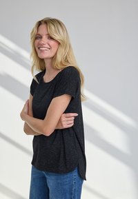 Blond naeratav naine käed risti, hõbedaste laikudega musta t-särgi ja siniste teksadega, seisab valge seina taustal varjuliste joontega.