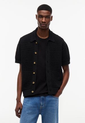 Νέος άνδρας που φοράει μαύρο πλεκτό κοντομάνικο ζακέτα με κουμπιά πάνω από μαύρο μπλουζάκι και μπλε τζιν, όρθιος με το ένα χέρι στην τσέπη.