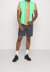 Grüner ärmelloser Steppweste über einem braunen T-Shirt, kombiniert mit grauen Shorts und schwarzen Schuhen. Mit Reißverschluss und Netzenseitenpaneelen.