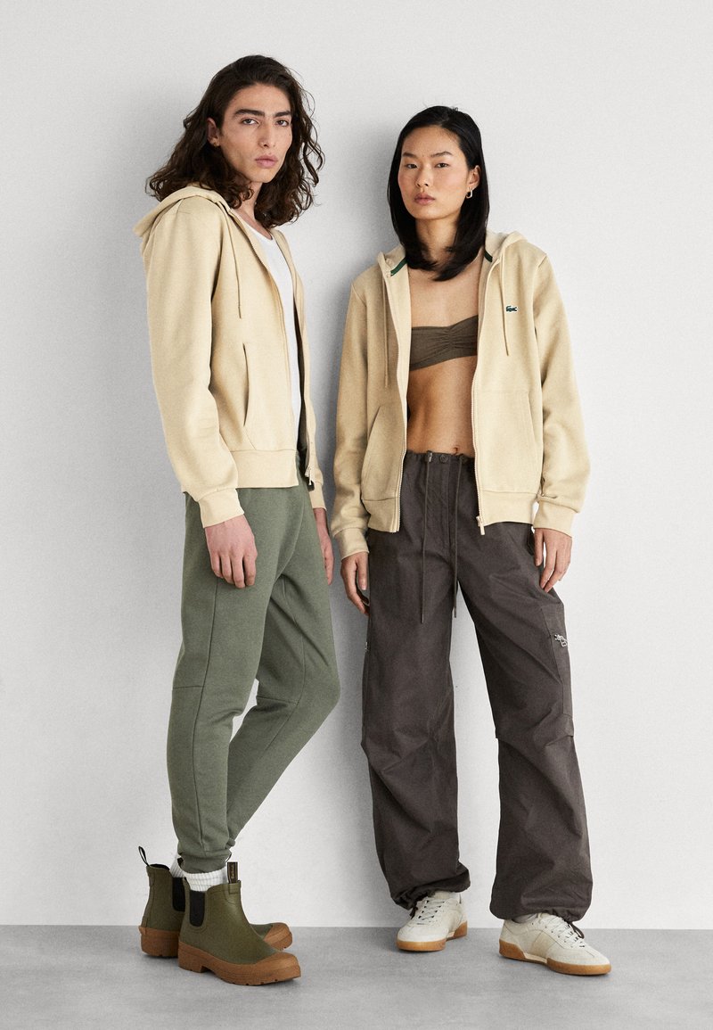 Beige ritsvest met groene accenten, olijfgroene joggers en donkergrijze cargo-broeken. Het mannelijke model draagt bruine laarzen; de vrouwelijke model heeft crèmekleurige sneakers aan.