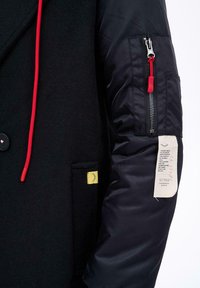 Černý kabát s červeným stahovacím šňůrkou, černým kapsou na rukávě na zip s červeným jezdcem a bílou štítkem s textem a malým grafickým motivem řasy.