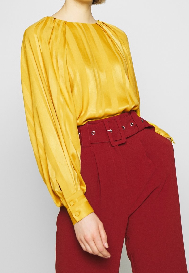 Blouse en satin jaune avec un col haut et des plis, associée à un pantalon taille haute bordeaux avec ceinture et œillets argentés.