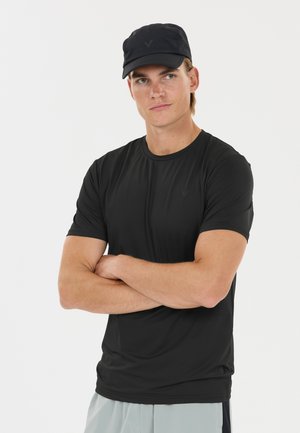 Young man with light skin wearing a black cap and black t-shirt, arms crossed, looking slightly to the side against a plain white background.
