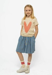 Niña pequeña con camiseta beige con un gran corazón rayado rojo, falda de cuadros azul, calcetas beige con rayas rojas y zapatillas blancas, sonriendo.