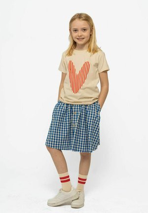 Jong meisje in een beige t-shirt met een grote rode gestreepte hart, een blauw geruit rokje, beige sokken met rode strepen en witte sneakers, lachend.