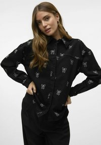 Zwarte shirt met witte strikkoorden, met knoopsluiting, lange mouwen en een klassieke kraag. De stof lijkt glad met een ontspannen pasvorm.
