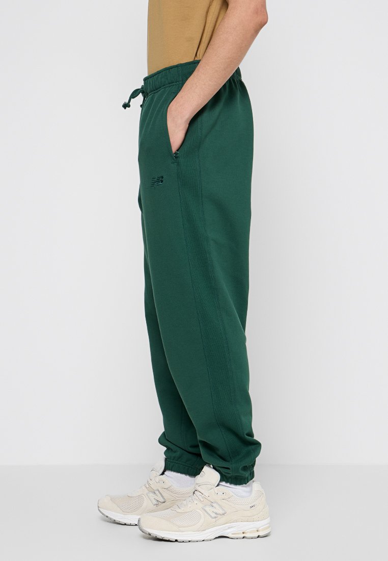 Pantaloni sportivi verdi con vita elastica e tasche laterali, caratterizzati da un design semplice e un logo sulla gamba. Indossati con sneaker beige.