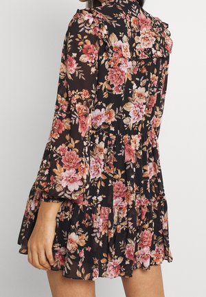 Kvinde iført en sort minikjole med lange ærmer, pink og orange blomsterprint samt flæsedetaljer på kant og skuldre.