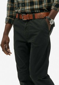 Homme portant un pantalon noir, une ceinture en cuir marron avec une étiquette, une chemise à carreaux avec les manches retroussées, et une montre noire avec un cadran argenté au poignet droit.