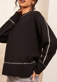 Maglione nero a maglia con orlo e polsini a coste, caratterizzato da cuciture bianche lungo le cuciture laterali. Tessuto testurizzato in un design loose.