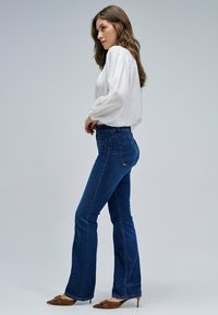 Vaqueros de mezclilla azul acampanados con una textura suave, combinados con una blusa blanca holgada. Zapatos de tacón marrones añaden contraste al atuendo.