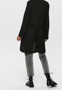 Czarna tunika z długim rękawem i wiązaniem w talii, wykonana z lekkiej tkaniny. Połączona z szarymi zwężanymi spodniami i czarnymi sznurowanymi botkami.