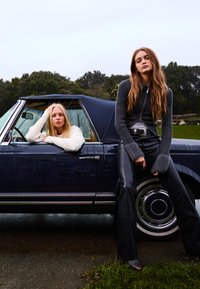 Två kvinnor poserar vid en marinblå cabriolet. Den ena lutar sig mot dörren i en vit kofta, medan den andra har på sig en grå topp och svarta läderbyxor.