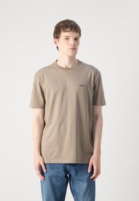 Kaki bomull T-skjorte med rund hals og korte ermer, med en liten svart "BOSS"-logo på venstre bryst. Brukt med blå jeans.