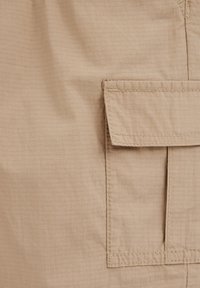 Tissu beige avec une poche à rabat cousue présentant une texture à motif quadrillé et des lignes de couture détaillées sur le côté droit.