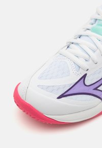 Hvit sportsko med teksturert overdel, lilla sidesymbol og rosa gummisåle. Inkluderer lyseblå detaljer og hvite snørebånd.