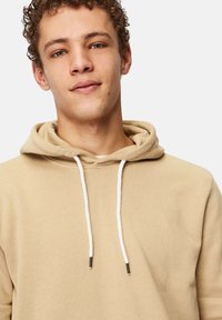 Beige Kapuzenpulli aus weichem Stoff, ausgestattet mit einer Kapuze mit einem Zugband in Weiß und metallenen Spitzen, präsentiert ein einfaches, sauberes Design.