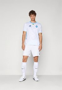 Valge jalgpallimärk siniste aktsentide ja logoga, koos valgete pükste ja sokkidega. Omaduseks on ümmargune krae ja mustriline materjal.