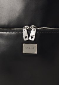 Zwart lederen oppervlak met twee zilveren ritsen van Guess boven een rechthoekige metalen Guess Los Angeles-logoplaat.