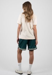 Femme aux cheveux blonds ondulés portant un t-shirt beige, un short de sport vert avec une bordure beige, des chaussettes blanches et des mocassins beiges, debout devant un fond uni.