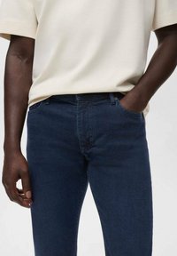 Persona vestida con jeans azul oscuro y una camisa beige de manga corta, con la mano izquierda en el bolsillo y la mano derecha relajada a un lado.