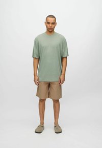 Camiseta de manga corta verde claro, pantalones cortos beige y zapatos gris de tipo slip-on, de pie contra un fondo liso, paleta de colores neutros.