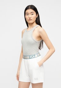 Jeune femme aux longs cheveux noirs portant un haut gris sans manches et un short blanc avec le logo "EA7", debout avec une main dans la poche.