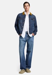 Giacca di jeans con colletto in camoscio marrone, chiusura con bottoni, due tasche sul petto, indossata su una camicia bianca e jeans blu a gamba larga. Scarpe casual.