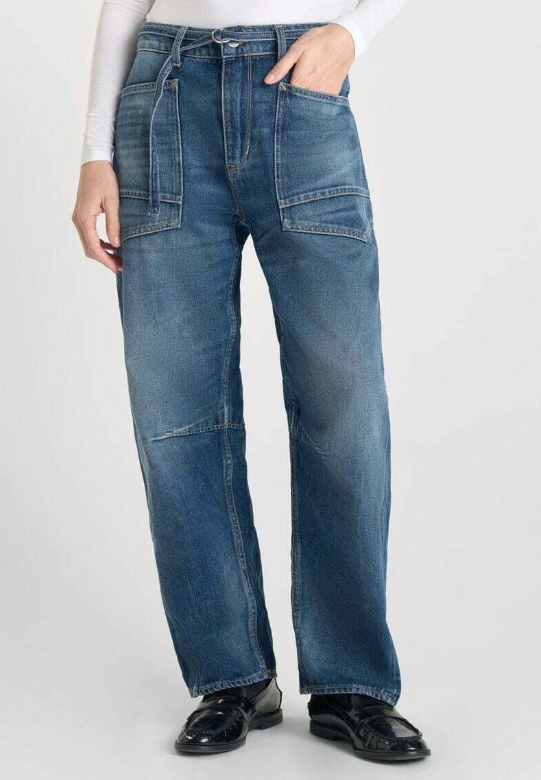Personne portant un jean bleu taille haute avec de grandes poches frontales et une ceinture en tissu, associé à des mocassins noirs et un haut à manches longues blanc.