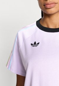 Svetlo vijolična majica s črnim ovratnikom in vezenim logotipom. Dizajn vključuje večbarvne črte na ramenih. Mehko blago.