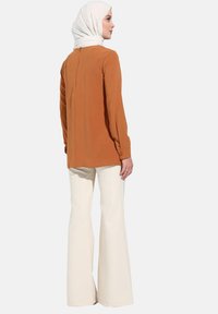 Blouse marron �à manches longues avec une fente dans le dos, assortie à un pantalon blanc évasé. La tenue présente un design minimaliste et des textures lisses.