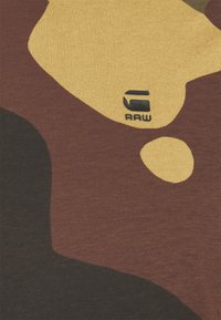 T-shirt med ett kamouflage-mönster i brunt, gult och svart. Mjuk tygkvalitet med en liten svart logotypdetalj nära mitten.