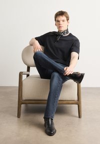 Černé tričko s krátkým rukávem, tmavé džíny, černé kožené kotníkové boty a vzorovaný černý šátek, sedící na moderní béžové židli.
