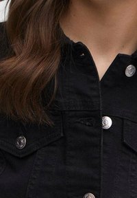 Giacca di denim nera con bottoni automatici argentati, collo appuntito e cuciture visibili. La texture appare liscia con una finitura leggermente opaca.