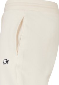 Joggers crema con cintura acanalada, con un bolsillo lateral y un parche de logo de estrella negra en la parte inferior de la pierna izquierda. Textura de tela suave.