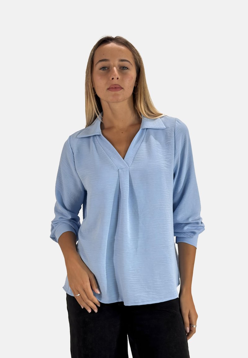 Blouse bleu clair à manches longues avec un col en V et des plis, présentant un tissu texturé et une coupe décontractée. Portée avec un pantalon noir.