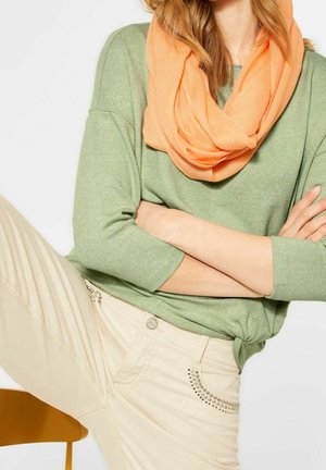 Vrouw met lichtgroene trui, perziksjaal, crèmekleurige broek met strassteendetails, armen gekruist en één been op een stoel geplaatst.