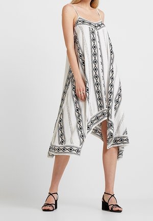 Kvinde iført hvid ærmeløs midi-kjole med sorte geometriske mønstre og sorte sandaler med blokhæl med stropper, stående mod en ensfarvet baggrund.