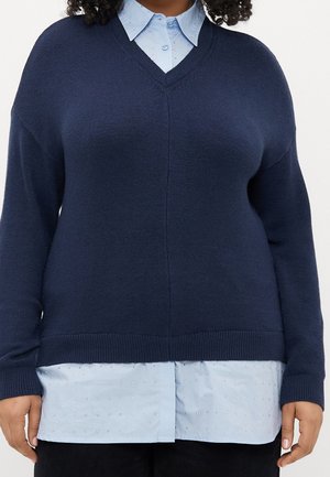 Vrouw draagt een marineblauwe V-hals trui over een lichtblauw overhemd met textuurelementen, zwarte broek, staand tegen een witte achtergrond.