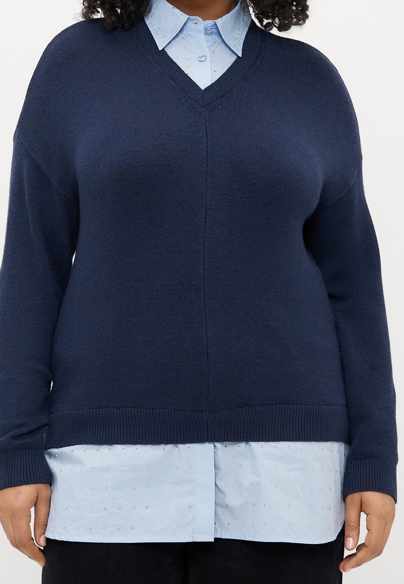 Femme portant un pull col en V bleu marine sur une chemise bleue claire à boutons avec des points texturés, pantalon noir, debout devant un fond blanc.