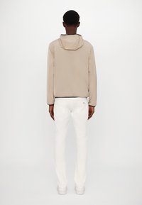 Beige, leichte Jacke mit Kapuze, schwarzem Saum und glatter Textur. Getragen mit enganliegenden weißen Hosen und weißen Schuhen.